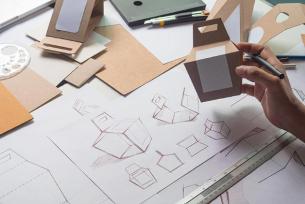 Hand sketching packaging concepts beside cardboard mock-ups